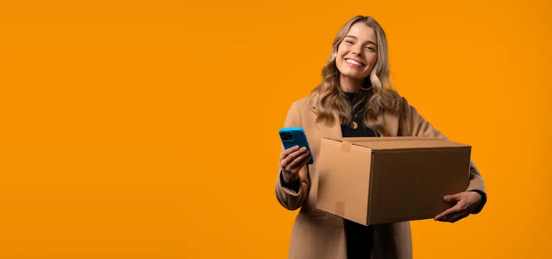 Lächelnde Frau hält ein Paket und ihr Smartphone vor einer orangefarbenen Wand