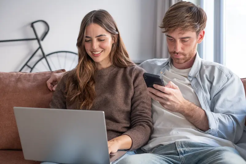 Paar sitzt auf dem Sofa und kauft mit Laptop und Smartphone online ein